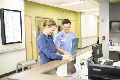 Two registered nurses looking at a chart at a reception desk in a hospital.