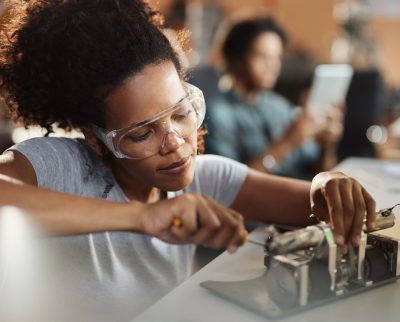 A woman wearing safety goggles using a screwdriver on a piece of hardware.